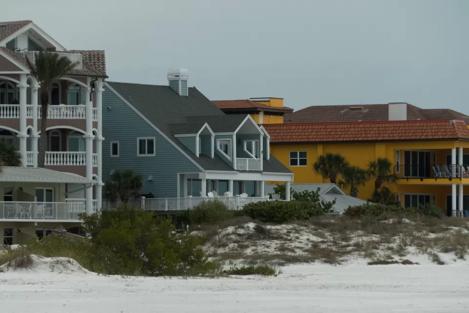 Three colorful, large beachfront homes, one khaki, one blue, and one yellow, with a small dune in front and flat sand in foreground.